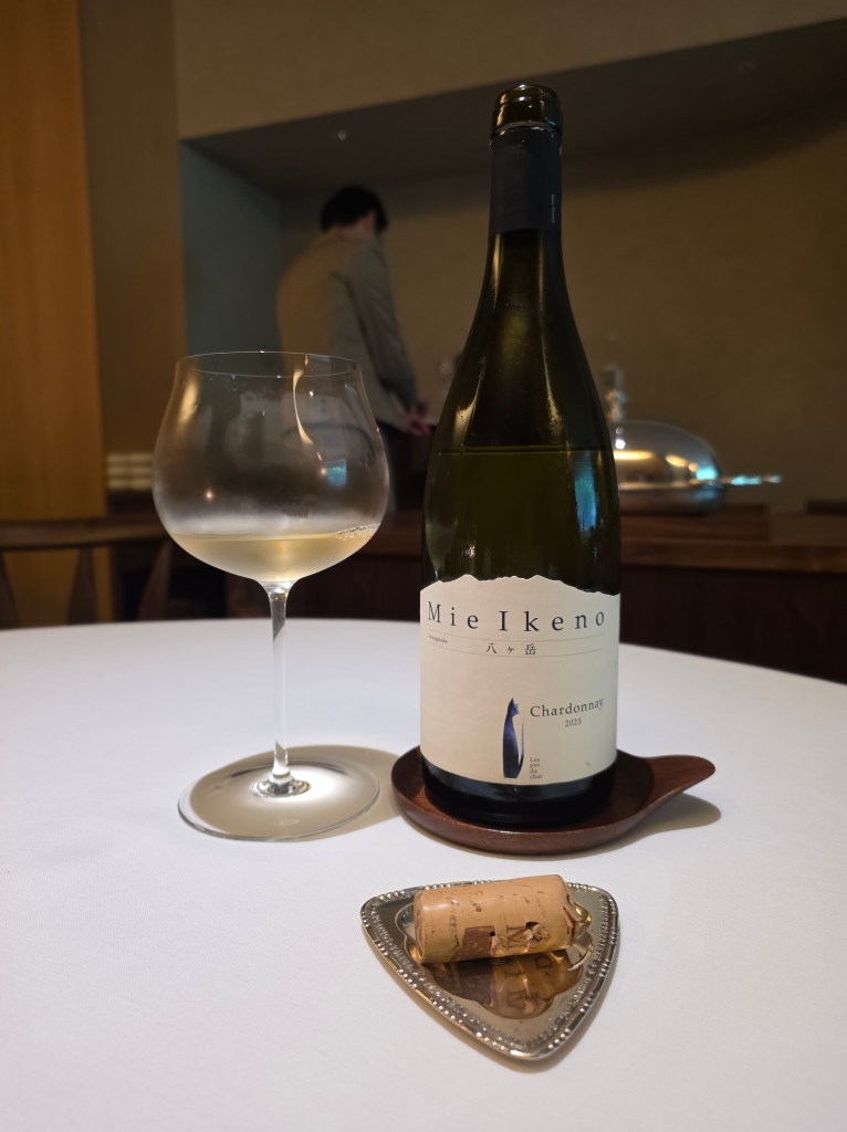 A bottle of Mie Ikeno Chardonnay 2023 next to a filled wine glass on a white tablecloth, with a wine cork on a silver dish in the foreground.