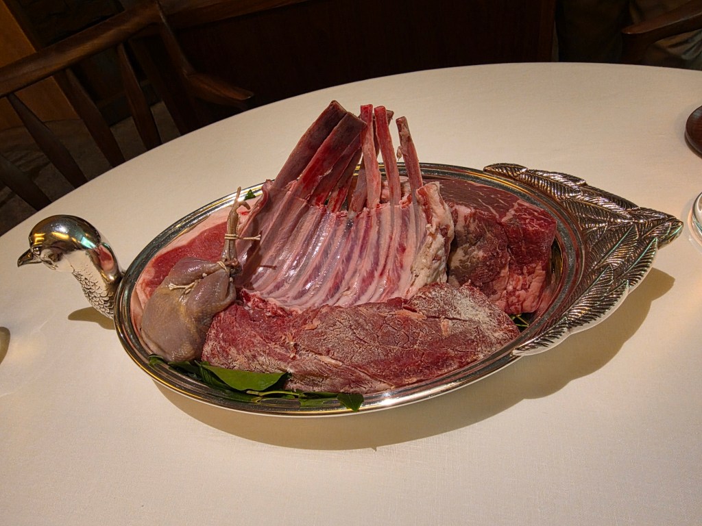A silver platter adorned with various cuts of raw meat, including a rack of lamb with exposed rib bones, alongside other meat cuts and garnished with green leaves.