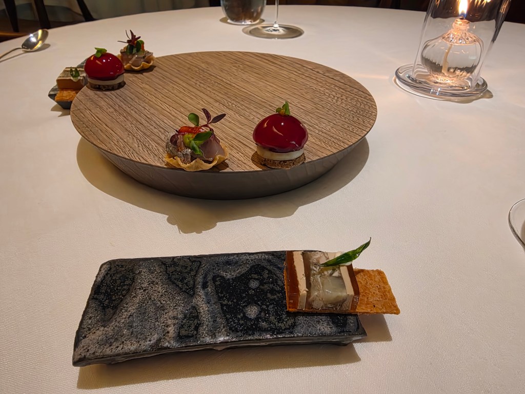 An elegantly presented dish featuring several gourmet appetizers on a wooden platter and a black stone plate. The appetizers are decorated with fresh herbs and colorful garnishes, set against a white tablecloth with a soft candle glow in the background.