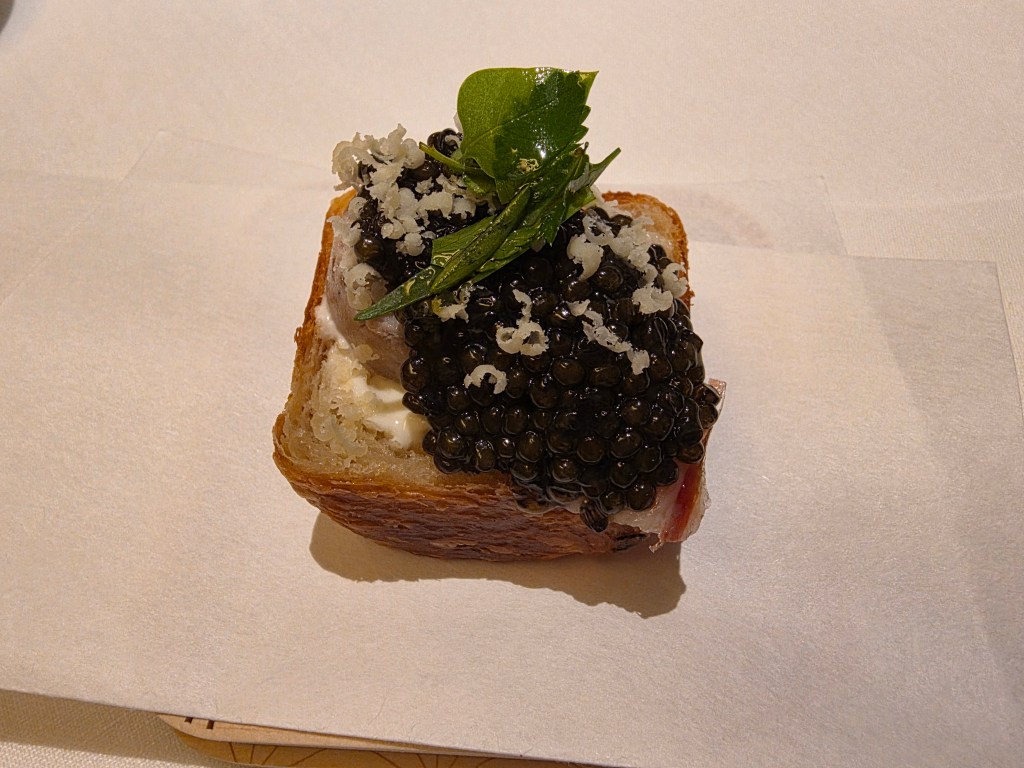 A small piece of toasted bread topped with a layer of caviar, white cream, and garnished with a green leaf and small flakes.