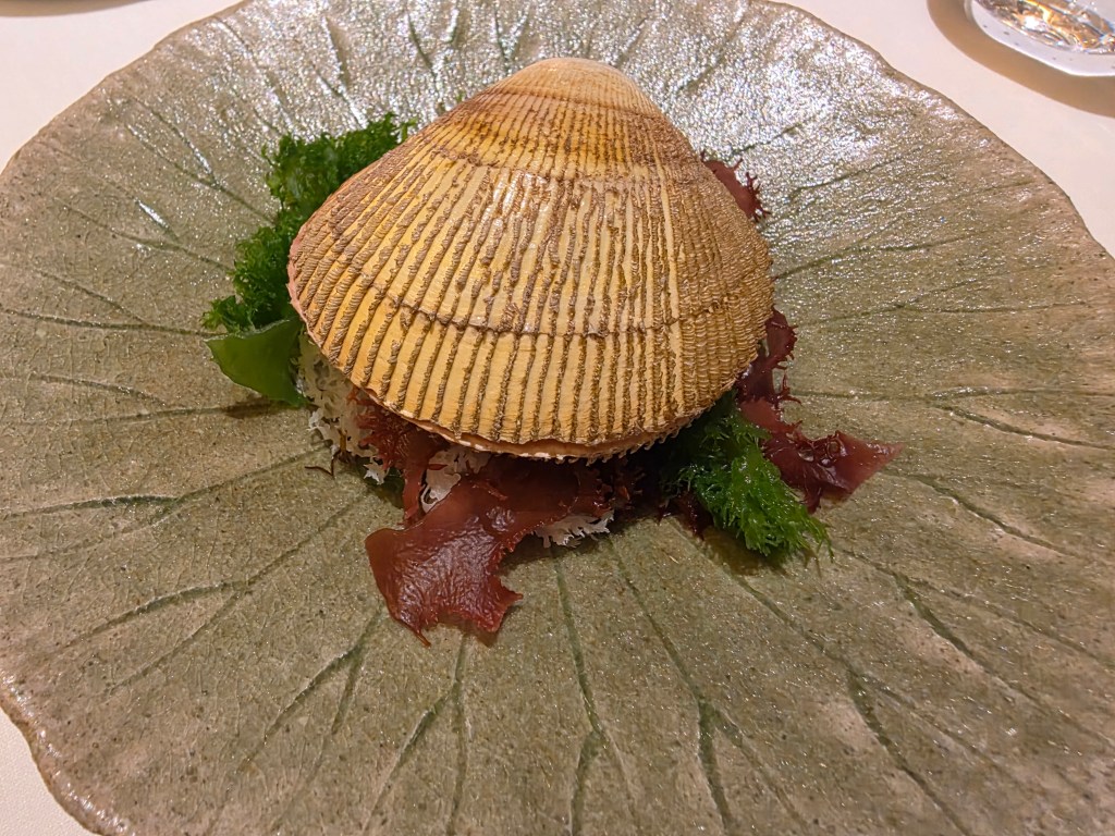 A large clam shell placed on a decorative green plate, surrounded by garnishes of seaweed and lettuce.