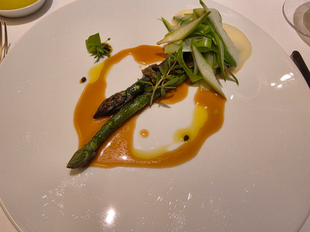 A plated dish featuring a single asparagus spear topped with garnishes, accompanied by a drizzle of sauce and thinly sliced greens.