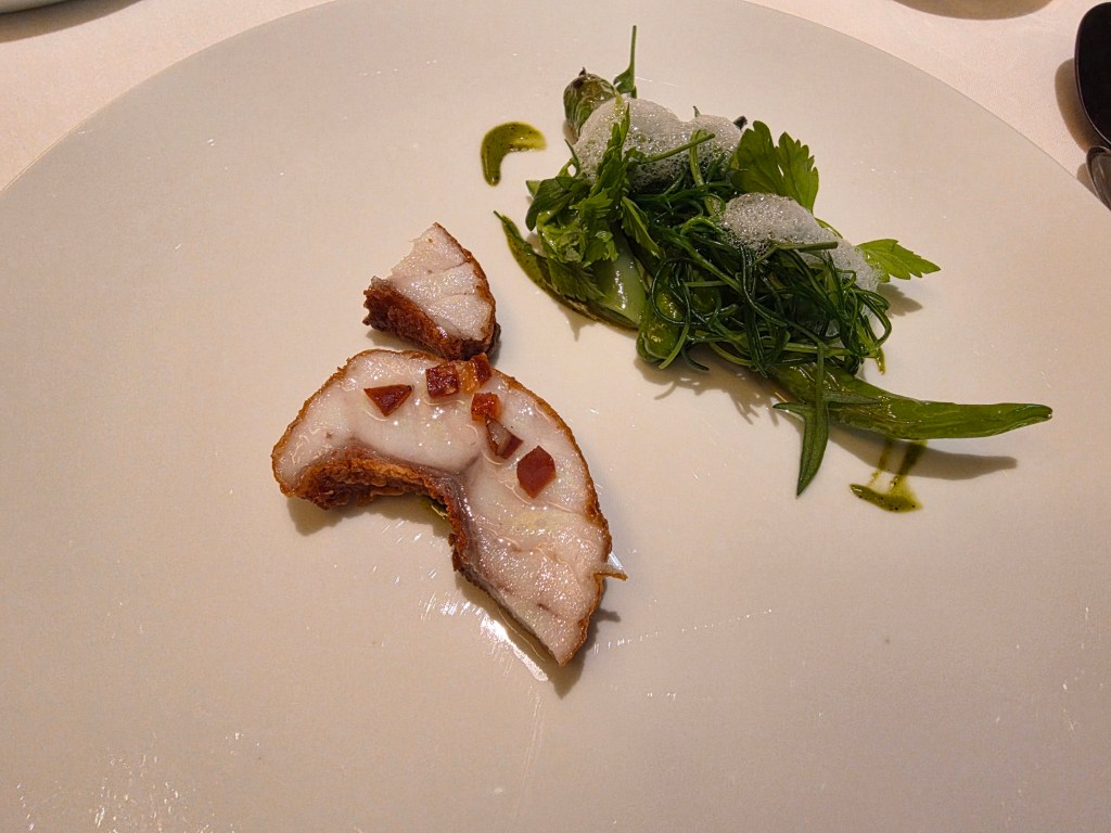 A plate featuring a slice of crispy pork belly topped with small pieces of red seasoning, accompanied by a side of fresh green herbs and edible foam.