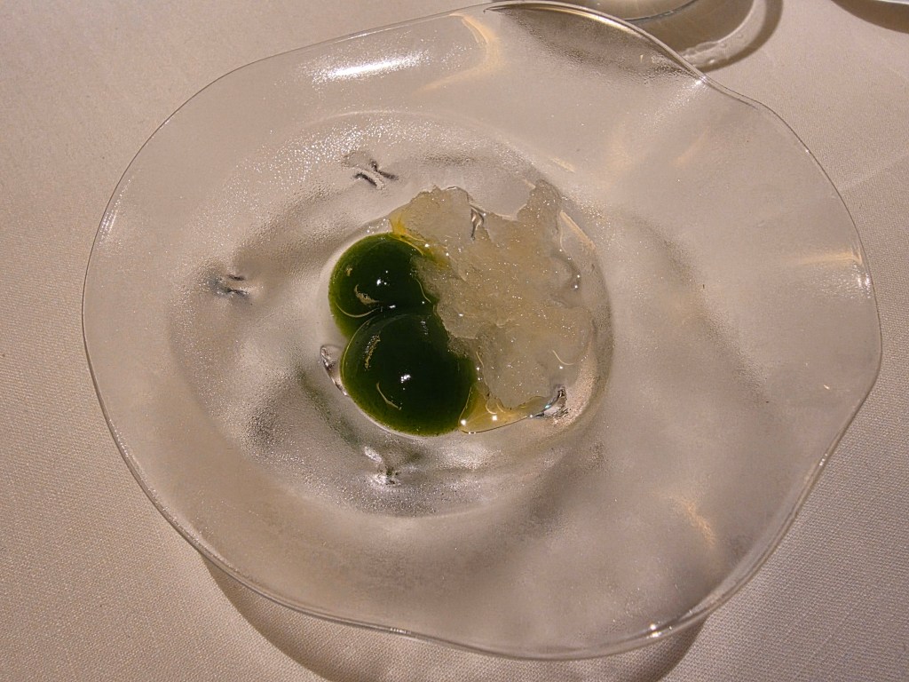 A delicate dish featuring two green spheres and a crystalline garnish on a clear, textured plate.