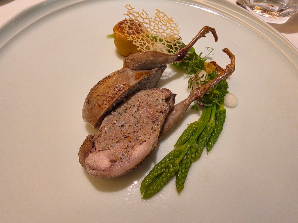A plated gourmet dish featuring roasted quail, sliced open to reveal its meat, accompanied by green herbs and decorative food elements.