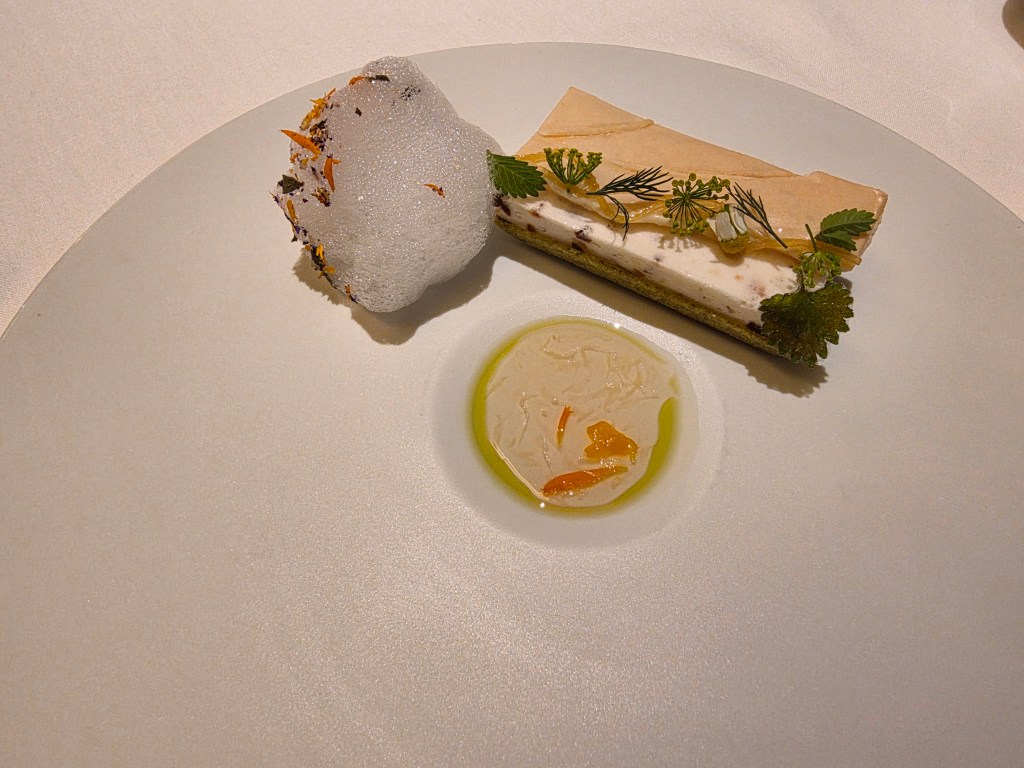 A gourmet dessert plate featuring a layered cake garnished with herbs, a piece of foamy confection, and a small serving of fruity sauce.