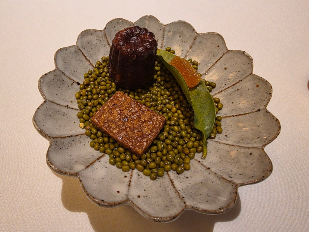 A decorative plate featuring green mung beans, a brown rectangular food item, a dark round dessert, and a green leaf with orange jelly.