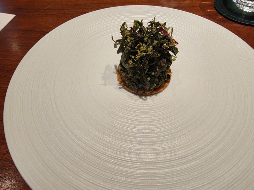 A gourmet dish featuring a small round pastry topped with an artful arrangement of green herbs and edible leaves, presented on a textured white plate.