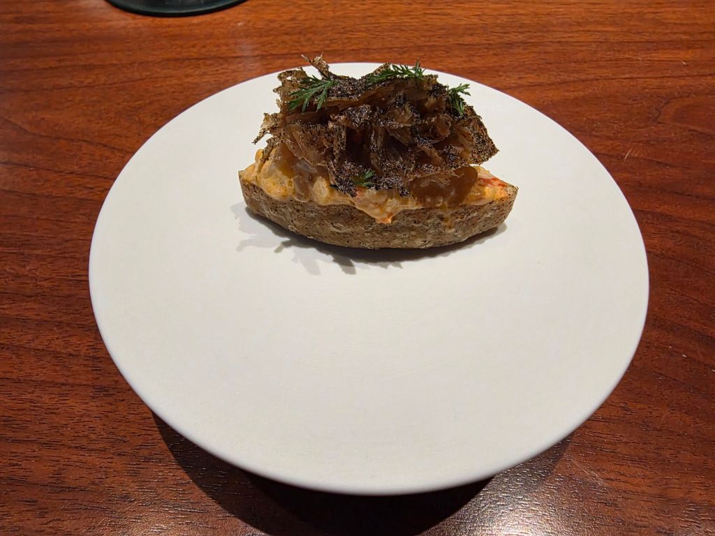 A slice of bread topped with a mixture of cream and herbs, garnished with crispy fried onions, served on a white plate.