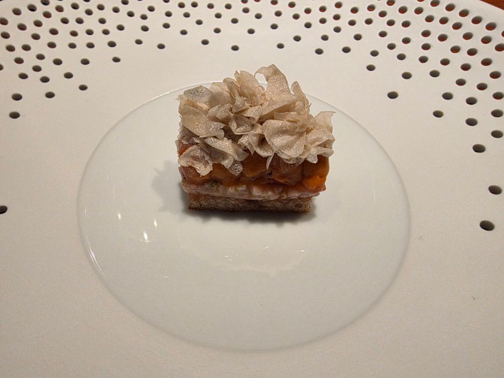 A small gourmet dish featuring a square piece of bread topped with colorful ingredients and delicate white edible decorations, presented on a minimalist plate.