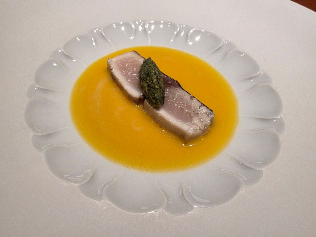 A gourmet dish featuring a slice of meat atop a vibrant yellow sauce, garnished with a dollop of green sauce, all on a white plate with a floral design.