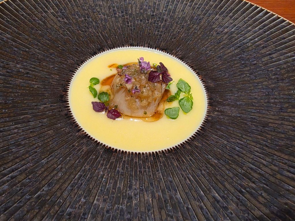 A gourmet dish featuring a piece of fish served on a creamy sauce, garnished with microgreens and edible flowers, presented on an intricately designed black plate.