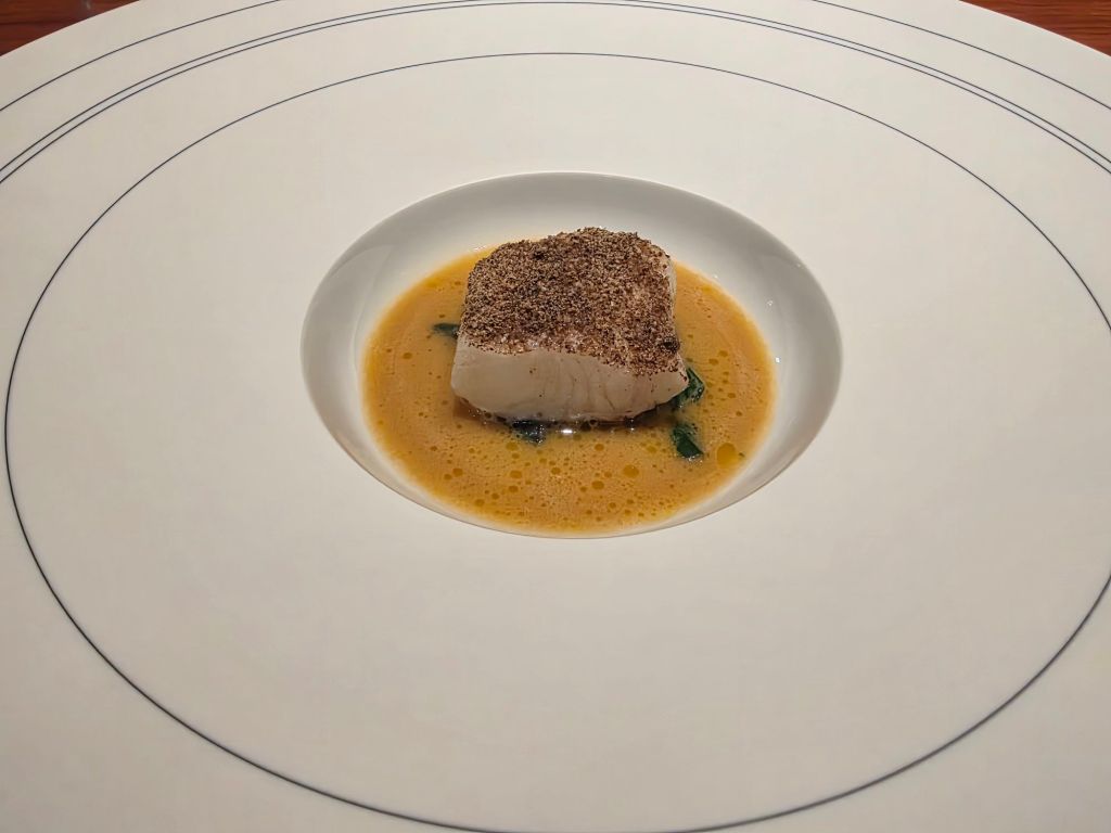 A plate featuring a piece of fish topped with a layer of spices, resting in a flavorful broth with green herbs, presented in an elegant white dish.