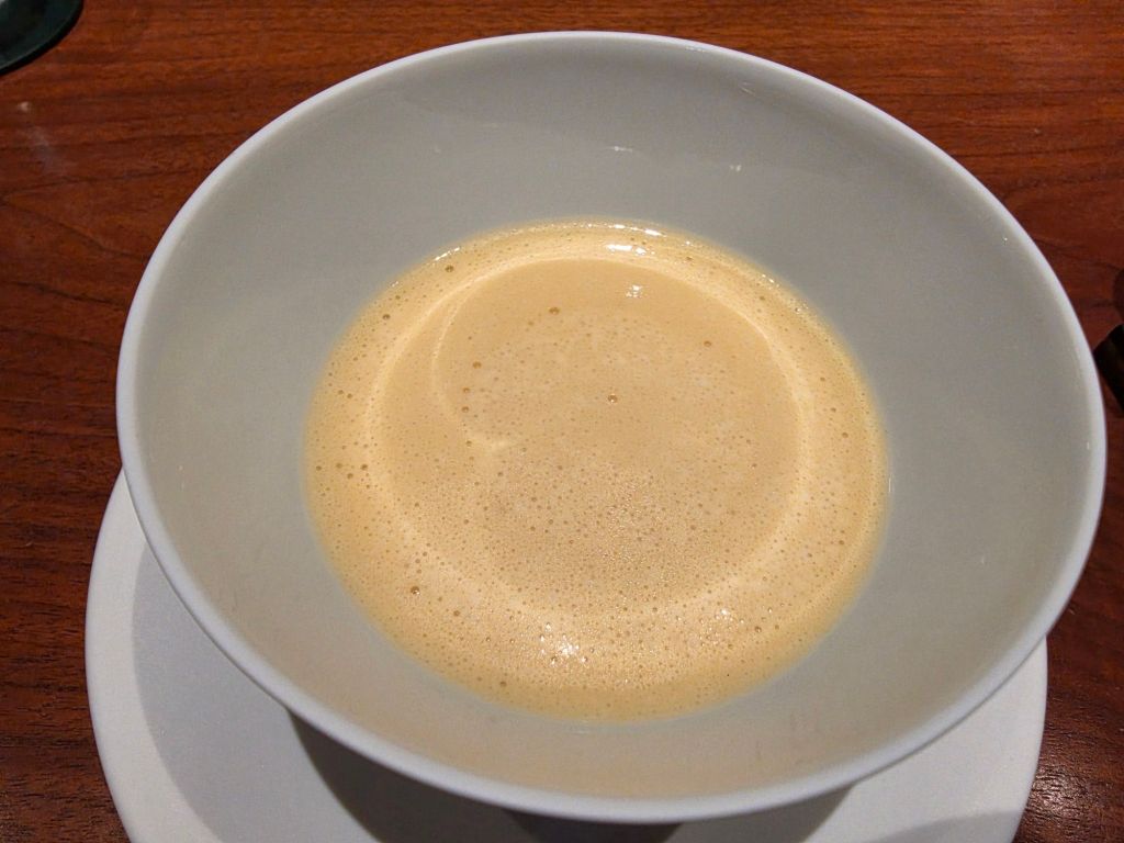A close-up view of a bowl containing a creamy, frothy coffee drink, resting on a wooden table.