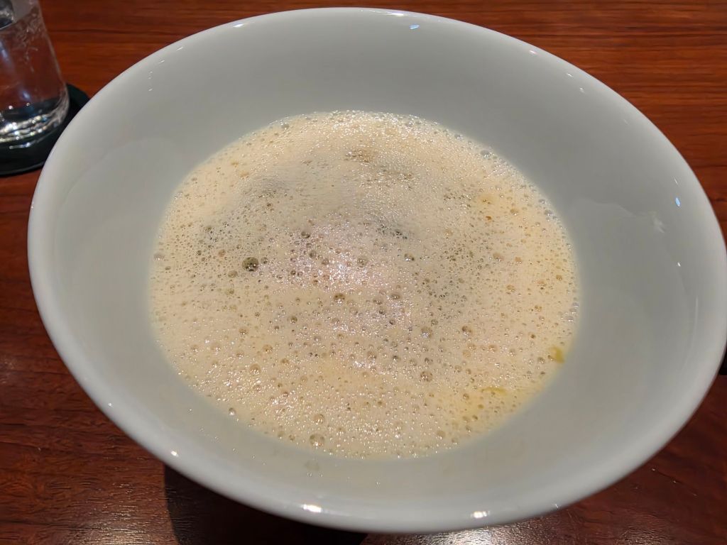 A white bowl filled with frothy liquid, possibly a broth or foam, placed on a wooden table.