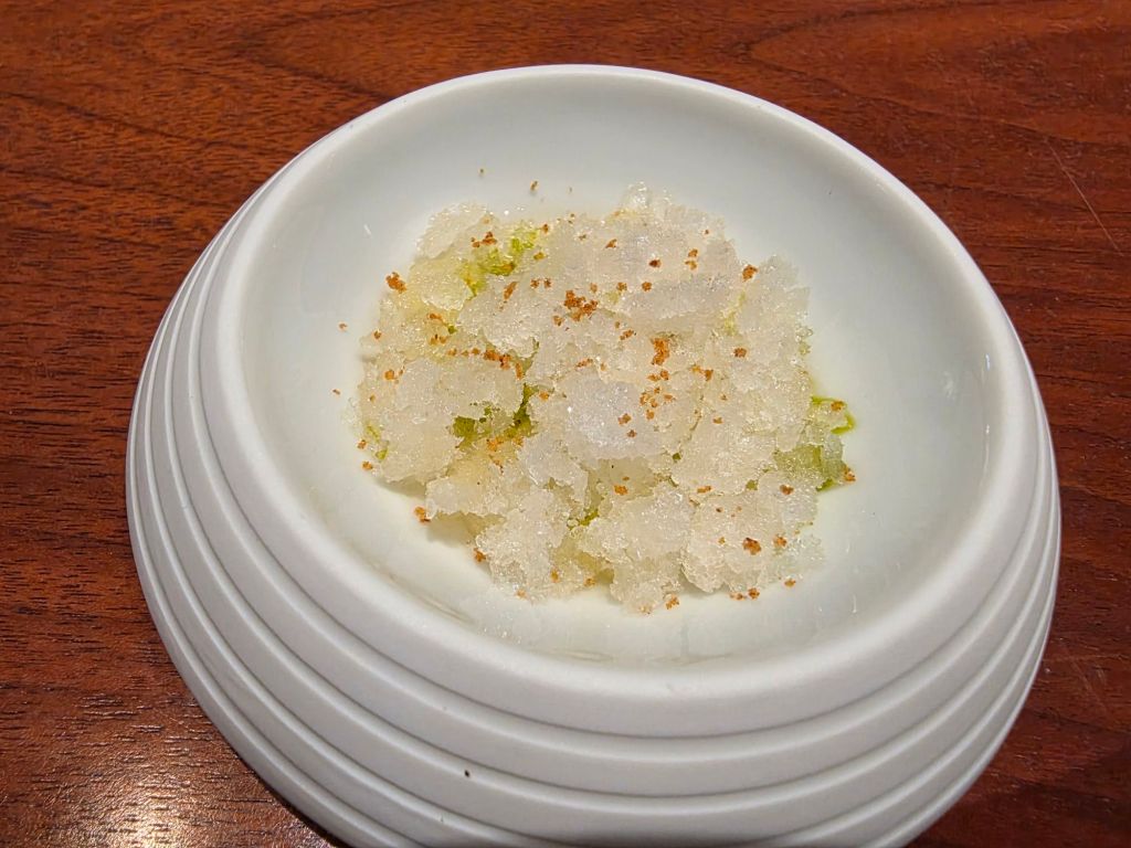 A small white dish filled with a textured, granular mixture resembling snow or crushed ice, lightly garnished with green and brown specks, resting on a wooden table.