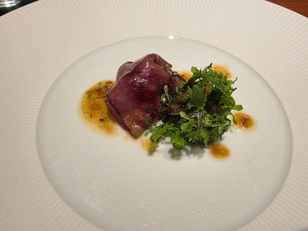 A plated dish featuring a piece of meat with a glossy sauce, accompanied by fresh green herbs on a white background.
