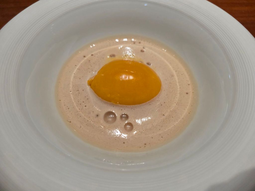 A plated dessert featuring a glossy yellow orb on a creamy, frothy brown sauce with small bubbles, presented in a white bowl.
