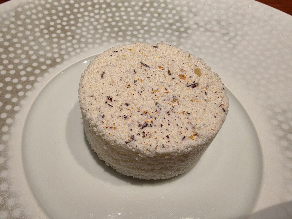 A round, white dessert with a textured surface, featuring specks of herbs and flowers, served on a white plate.