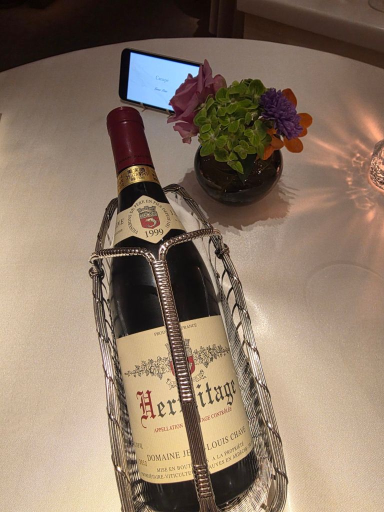A bottle of 1999 Hermitage wine in a silver wire holder on a table, accompanied by a small flower vase and a smartphone displaying a menu.