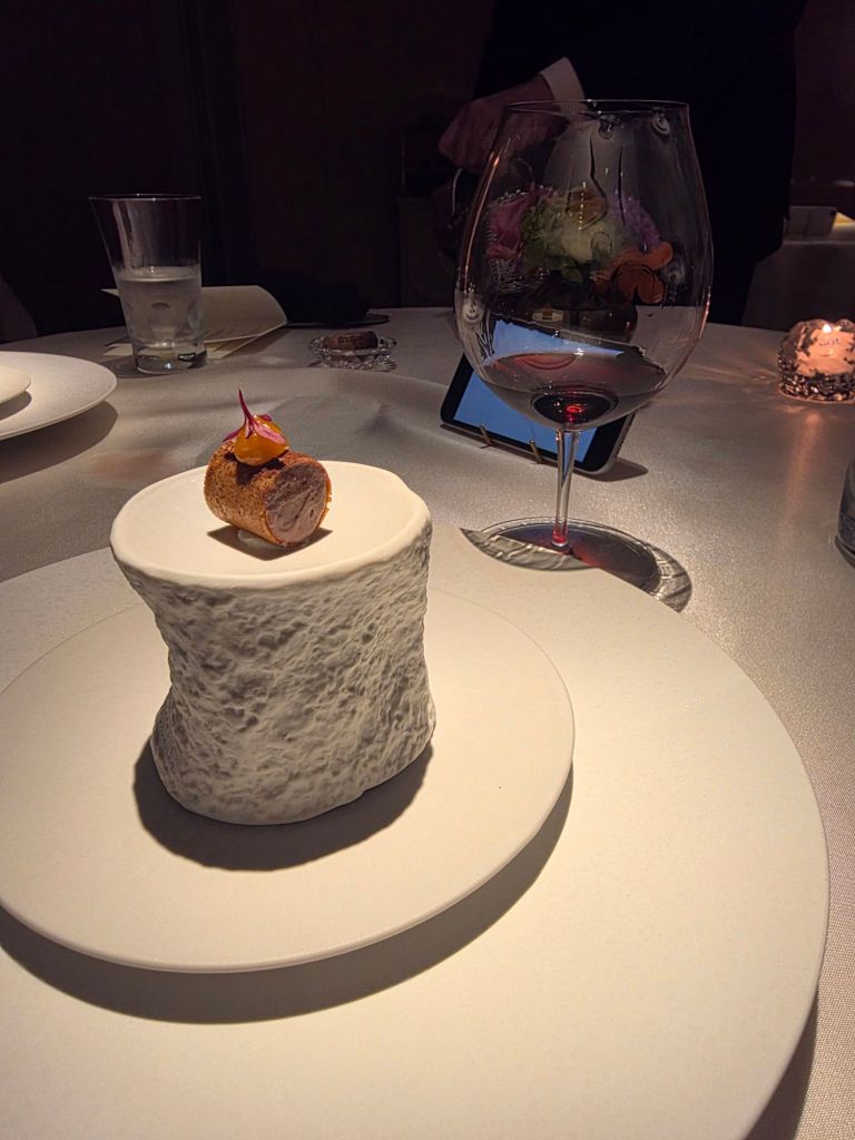 A gourmet dish featuring a textured white presentation plate with a unique shaped food item topped with a small garnish, accompanied by a wine glass and a water glass on a elegantly set table.