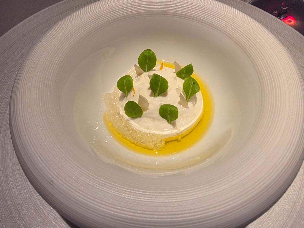 A gourmet dish featuring a creamy base topped with fresh green leaves, surrounded by a light oil drizzle and white foamy accents, presented in a sleek, round plate.
