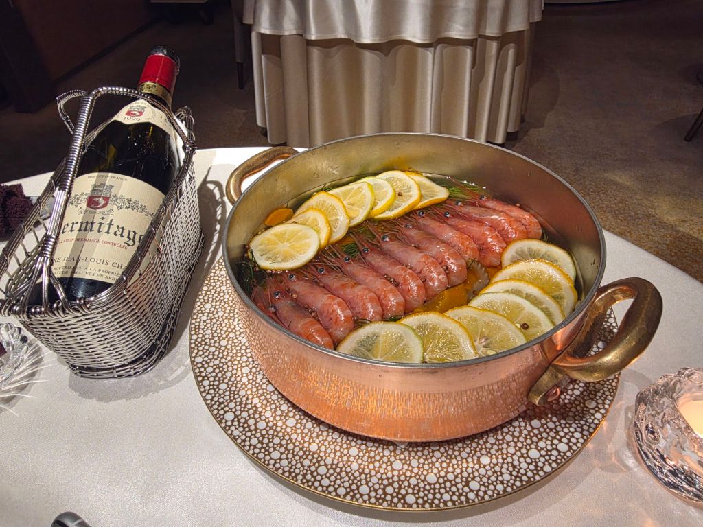 A copper pot filled with sliced fish and lemon garnishes, accompanied by a bottle of wine in a metal basket.