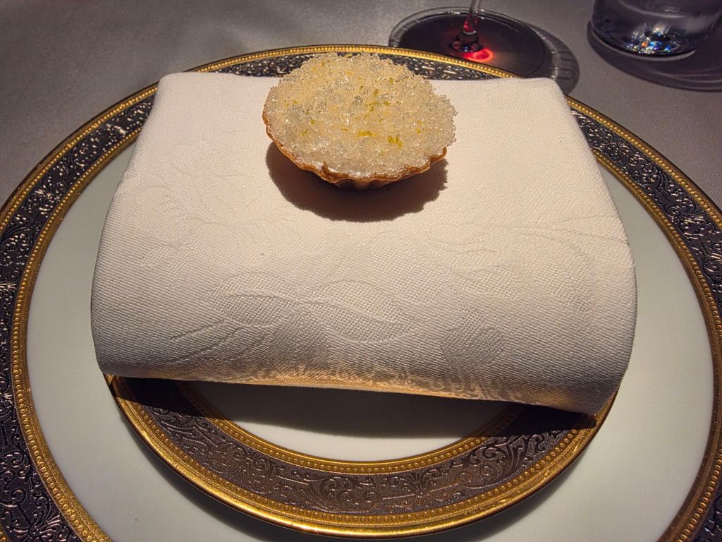 A elegant setting featuring a folded white napkin on a decorative plate, topped with a small food item resembling a mini tart with a crystalline texture.