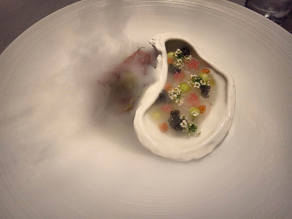 A beautifully presented dish featuring an oyster shell filled with colorful toppings, including various beads and garnishes, surrounded by a mist.