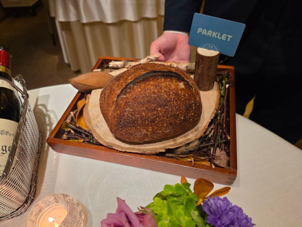 A rustic loaf of bread presented on a wooden platter with leaves and twigs, accompanied by a small sign labeled 'PARKLET'. A bottle of wine and a candle are nearby, along with colorful flowers.