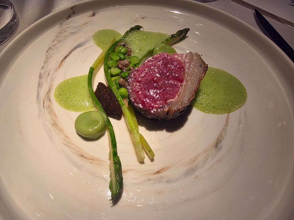 A gourmet dish featuring a beautifully presented piece of meat with a pink interior, surrounded by vibrant green vegetables and a delicate sauce.