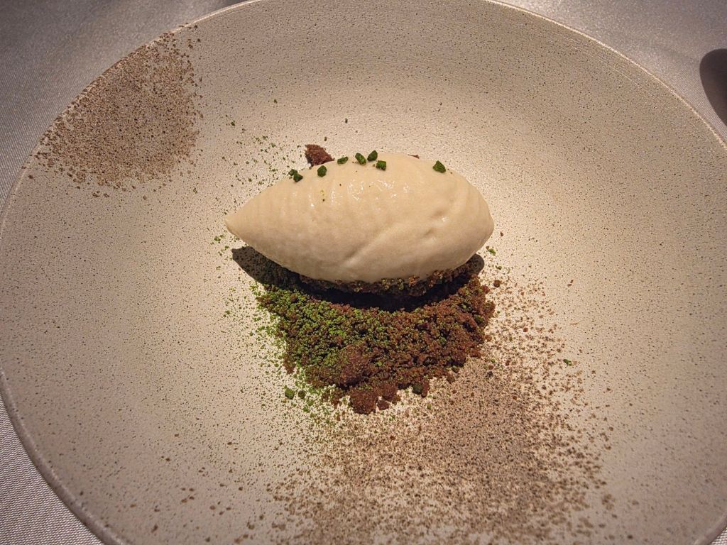 A minimalist dessert presentation featuring a creamy scoop of sorbet on a bed of finely crumbled chocolate and green herbal powder on a textured white plate.