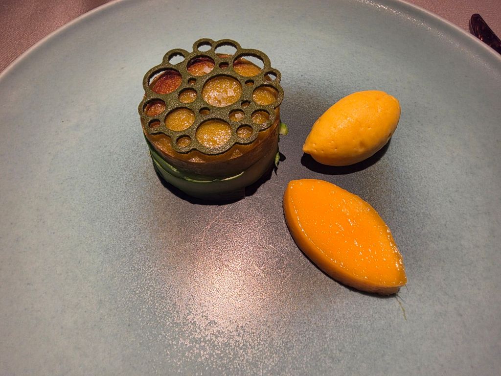 A beautifully presented dish featuring a circular green garnish atop a layered vegetable stack, accompanied by two yellow fruit sorbet pieces on a minimalist plate.