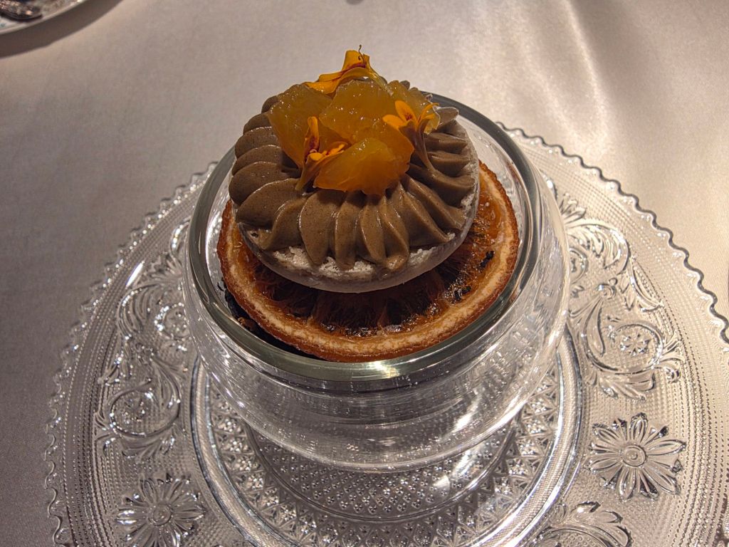 A gourmet dessert featuring a layered pastry topped with chocolate mousse, candied citrus pieces, and edible flowers, presented in a glass dish on an ornate plate.