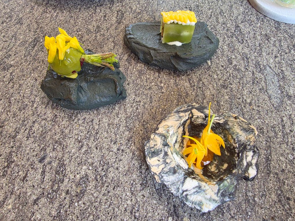 A creative presentation of three culinary items displayed on textured stone-like surfaces. The first item features a green round shape topped with yellow flowers, the second item is a small rectangular piece with green and white layers, and the third item resembles a natural bowl containing orange flowers.