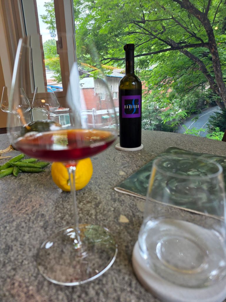 A close-up view of a glass of red wine on a countertop, with a bottle labeled 'RADIO' in the background, surrounded by a lemon and green peas, and a view of trees outside a window.