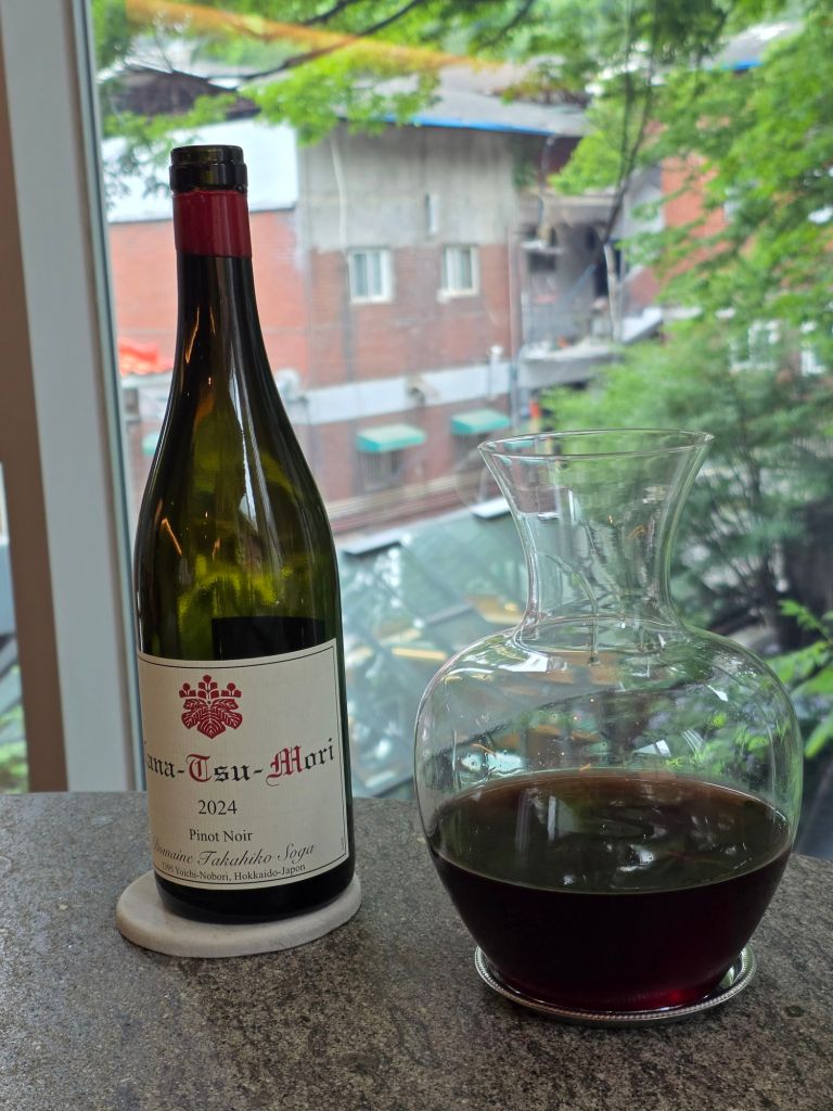 A bottle of 2024 Pinot Noir wine from Napa-Tsu-Mori next to a glass decanter filled with red wine, placed on a stone countertop with a view of greenery and buildings in the background.