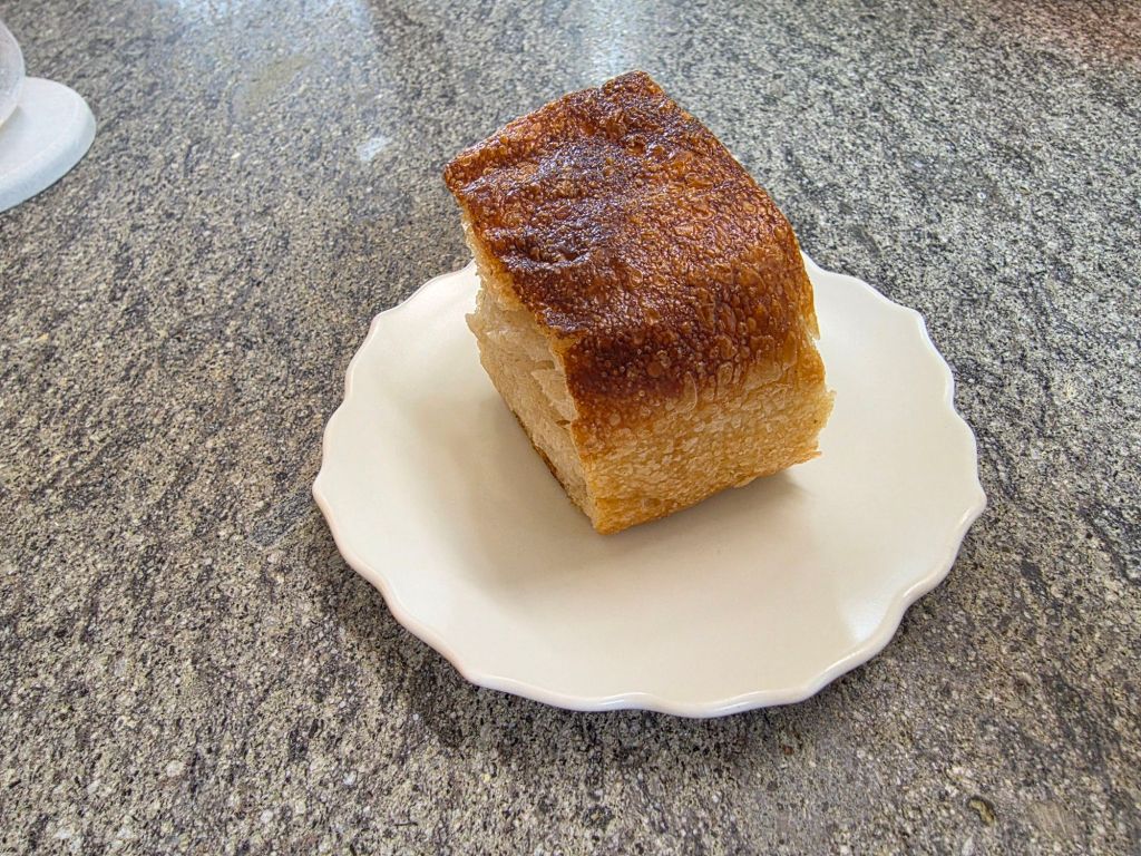 A slice of freshly baked bread with a golden, crispy crust on a white decorative plate, set against a textured gray surface.