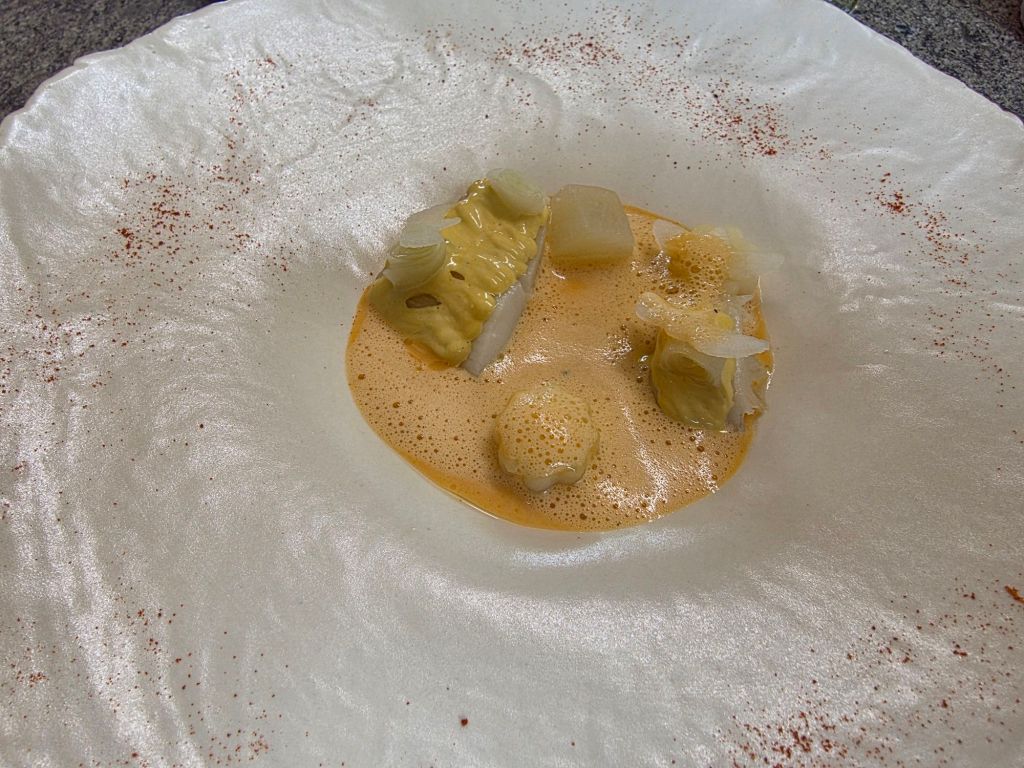 A gourmet dish presented on an elegant plate, featuring pieces of delicate fish and vegetables in a creamy sauce, garnished with light foam and a sprinkle of spice.