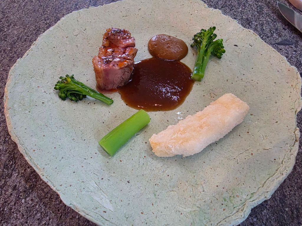 A gourmet dish served on a textured green plate, featuring a piece of seared meat, a sauce, cooked broccoli, a dollop of brown puree, and a white rectangular food item.