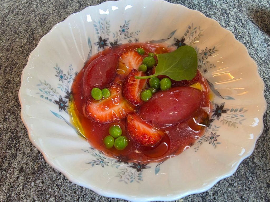 A decorative bowl filled with a colorful dessert featuring strawberry slices, green peas, and sorbet in a red syrup.