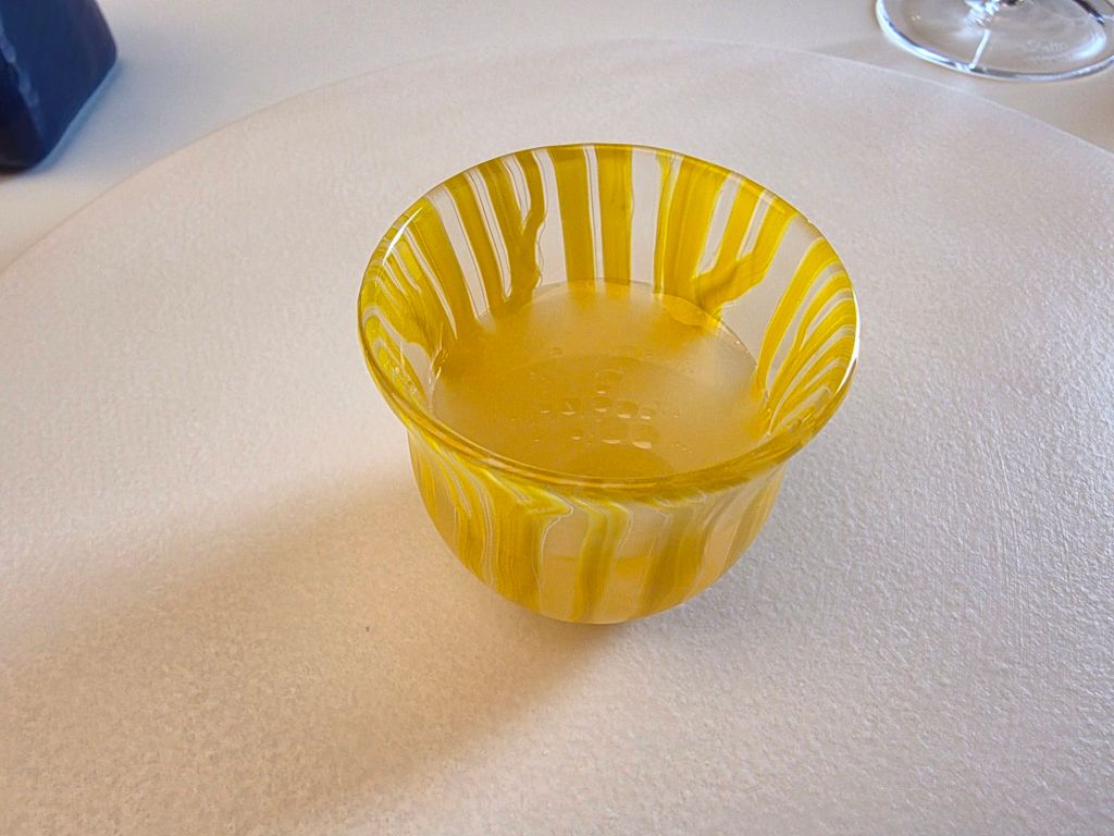 A small yellow glass bowl with a wavy striped pattern, containing a clear liquid, placed on a white surface.