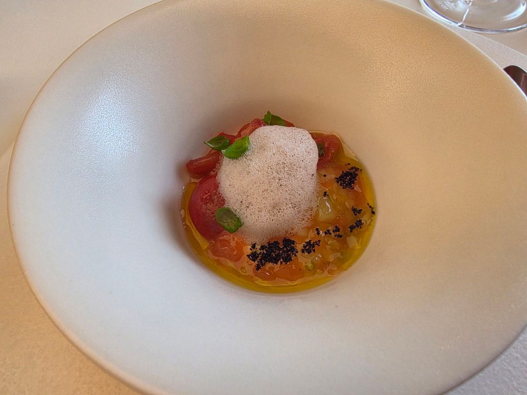 A gourmet dish presented in a white bowl, featuring a mix of colorful ingredients including small pieces of tomato, green herbs, and a light foam, surrounded by a drizzle of oil and sprinkled with black powder.