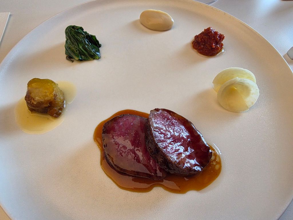 A plated dish featuring sliced meat with a glossy sauce, accompanied by various garnishes including a green leaf, dollops of creamy sauce, a piece of golden vegetable, and a red condiment on a white plate.