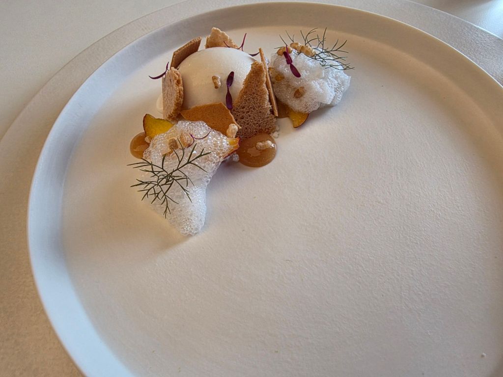 A gourmet dessert presented on a white plate, featuring a spherical white dessert surrounded by pieces of bread, colorful garnishes, and a light foam. Decorative elements include herbs and edible flowers.
