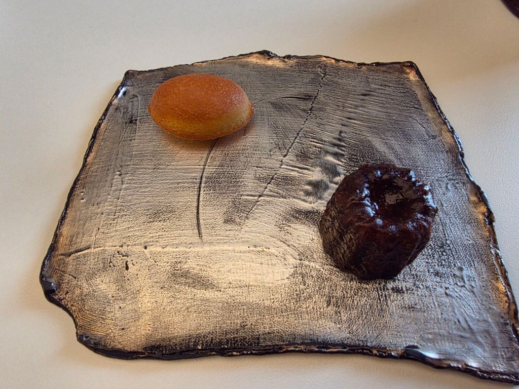 A gourmet dessert plate featuring a round, orange pastry and a small, dark-brown cake on a textured, metallic serving platter.