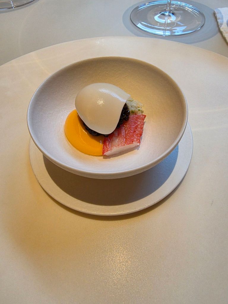 A gourmet dish served in a white bowl, featuring a poached egg, a dollop of sauce, pieces of crab meat, and a bright orange yolk placed on a circular plate.