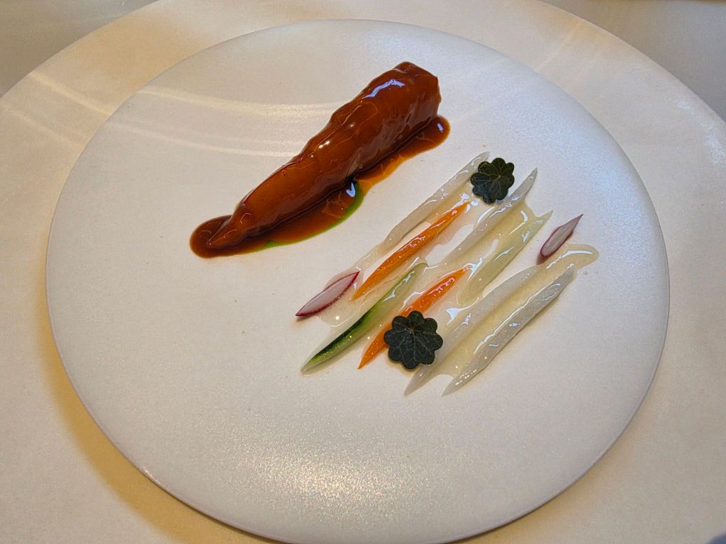 A gourmet dish featuring a glossy piece of protein drizzled with sauce, accompanied by neatly arranged strips of colorful vegetables and garnishes on a white plate.