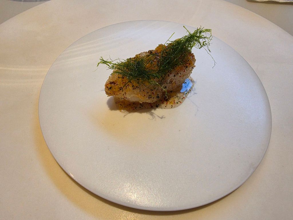 A gourmet dish featuring a small piece of seasoned fish topped with green herbs, presented on a minimalist white plate.