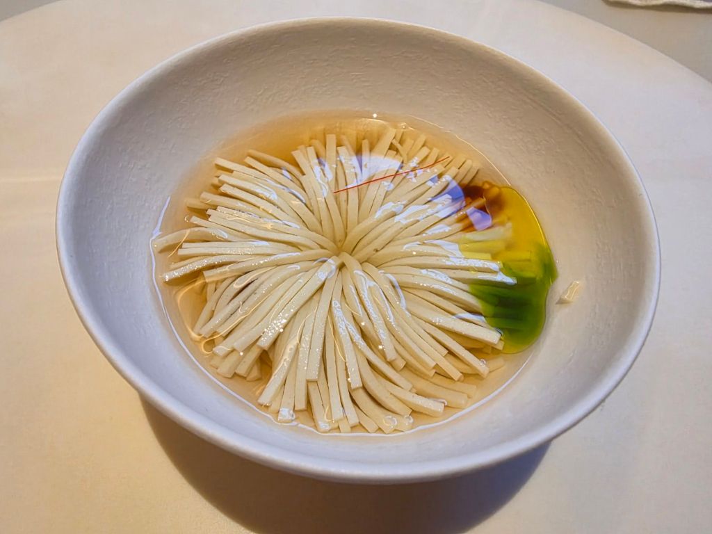 A bowl containing thin white noodles arranged in a floral pattern, submerged in a light, clear broth with a touch of green and an edible flower for garnish.